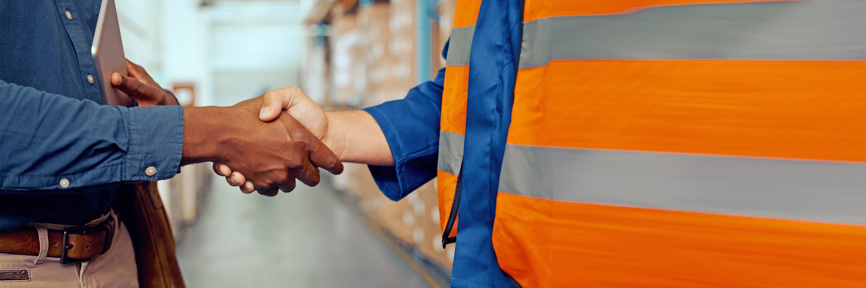 Two people shake hands as partners. The picture shows a close-up of only the two hands. The person on the left is wearing a blue shirt and the person on the right is wearing an orange safety vest. The surroundings of a workshop can be seen in the background.