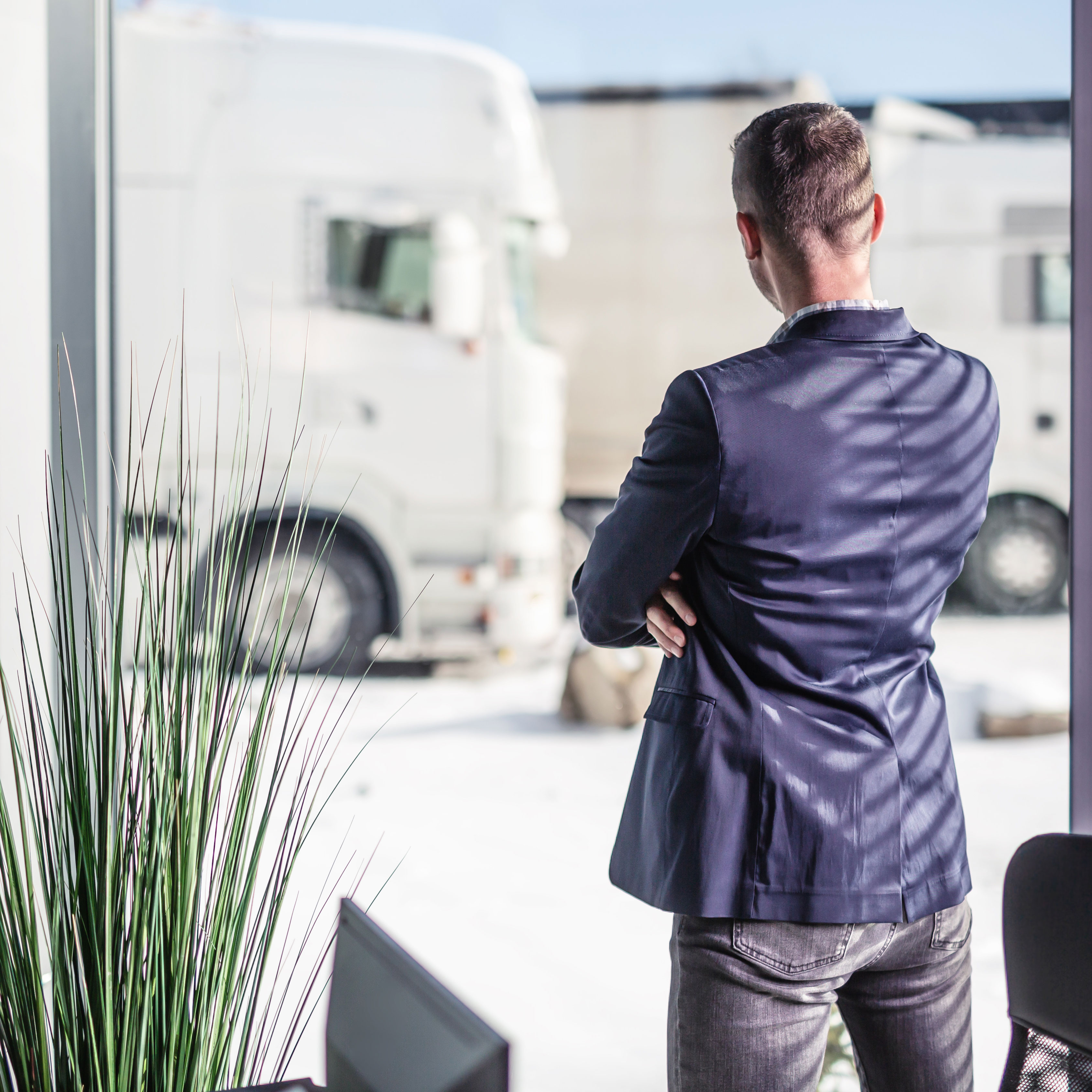 Picture of a fleet manager in a jacket and jeans, standing at the window and looking at his trucks, which can be seen vaguely in the background. 