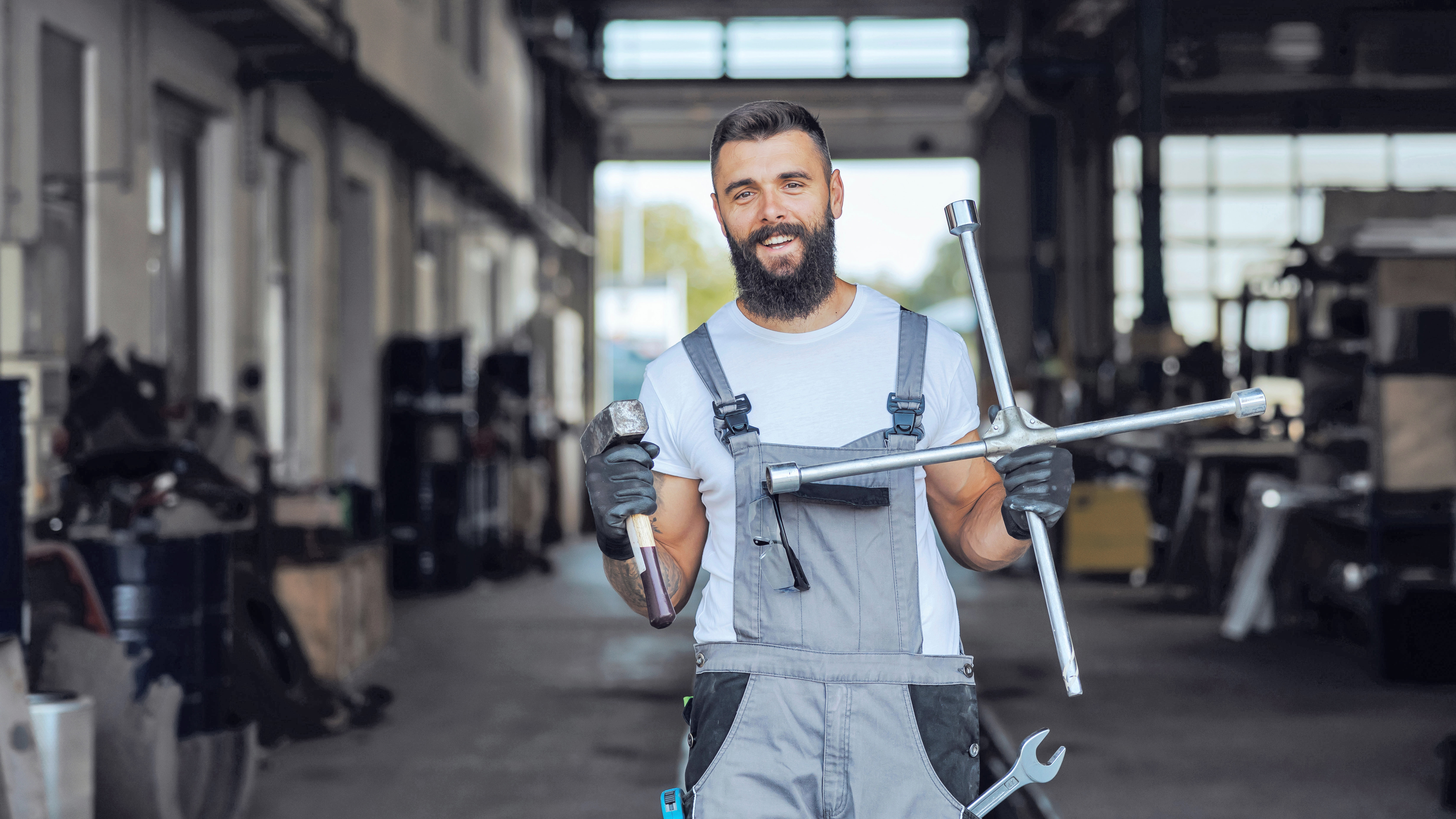 You can see a young man with a beard in typical workshop work clothes. A wrench protrudes from the gray overall and the workshop employee has a hammer and a cross wrench in his hands. A car repair shop can be seen slightly out of focus in the background.